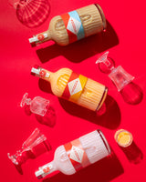 Three bottles of Mizzica liqueur with colorful labels on a red background, surrounded by wine glasses and a small glass container.